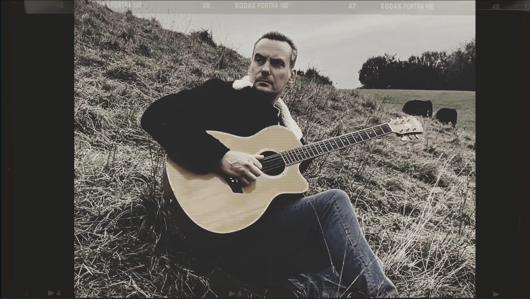 Sam Brandon seated outdoors with an acoustic guitar while filming the Come Home music video